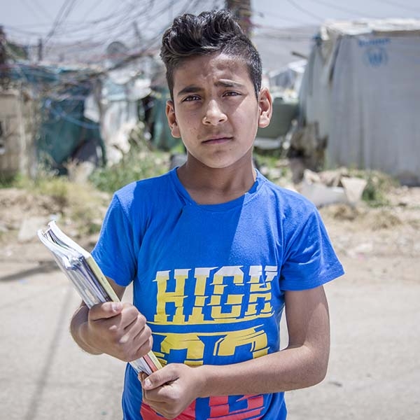 Siraj, age 13, looks concerned, as he stands in the informal, tented settlement where he lives, holding some notebooks.