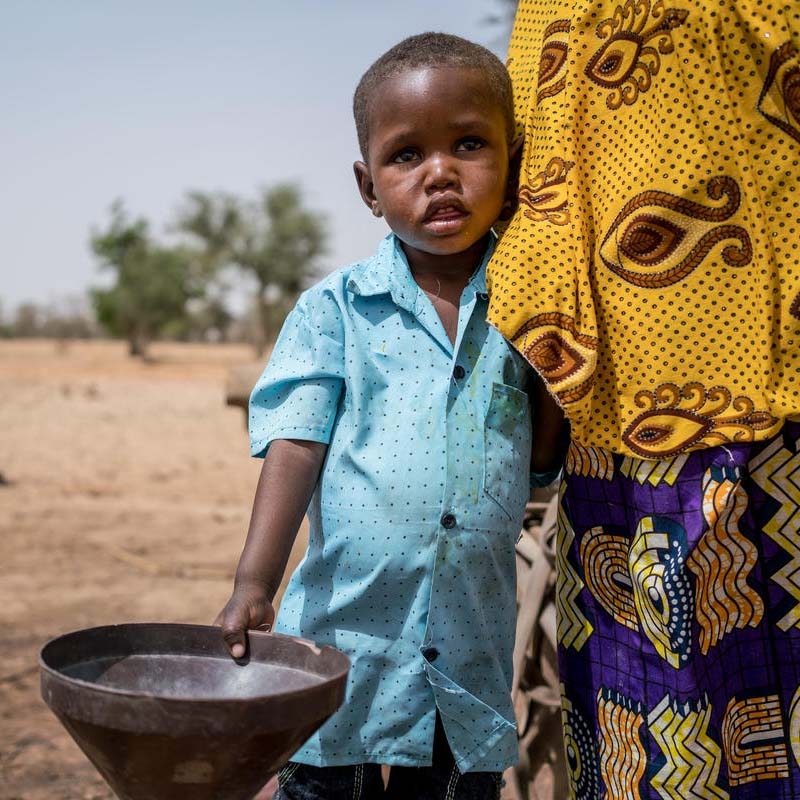In Nigeria, a young boy hugs his mother's leg while standing outside and holding a bucket.