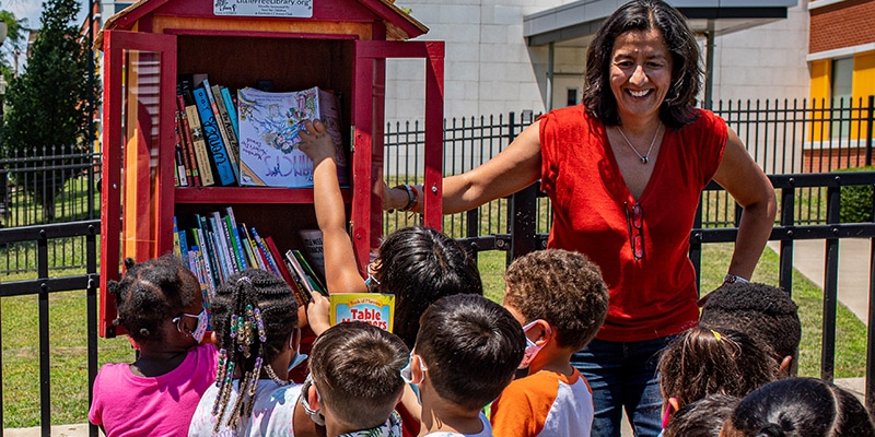 Premiumaid Foundation CEO Janti Soeripto stands near a Little Free Library while children enjoy book selection.