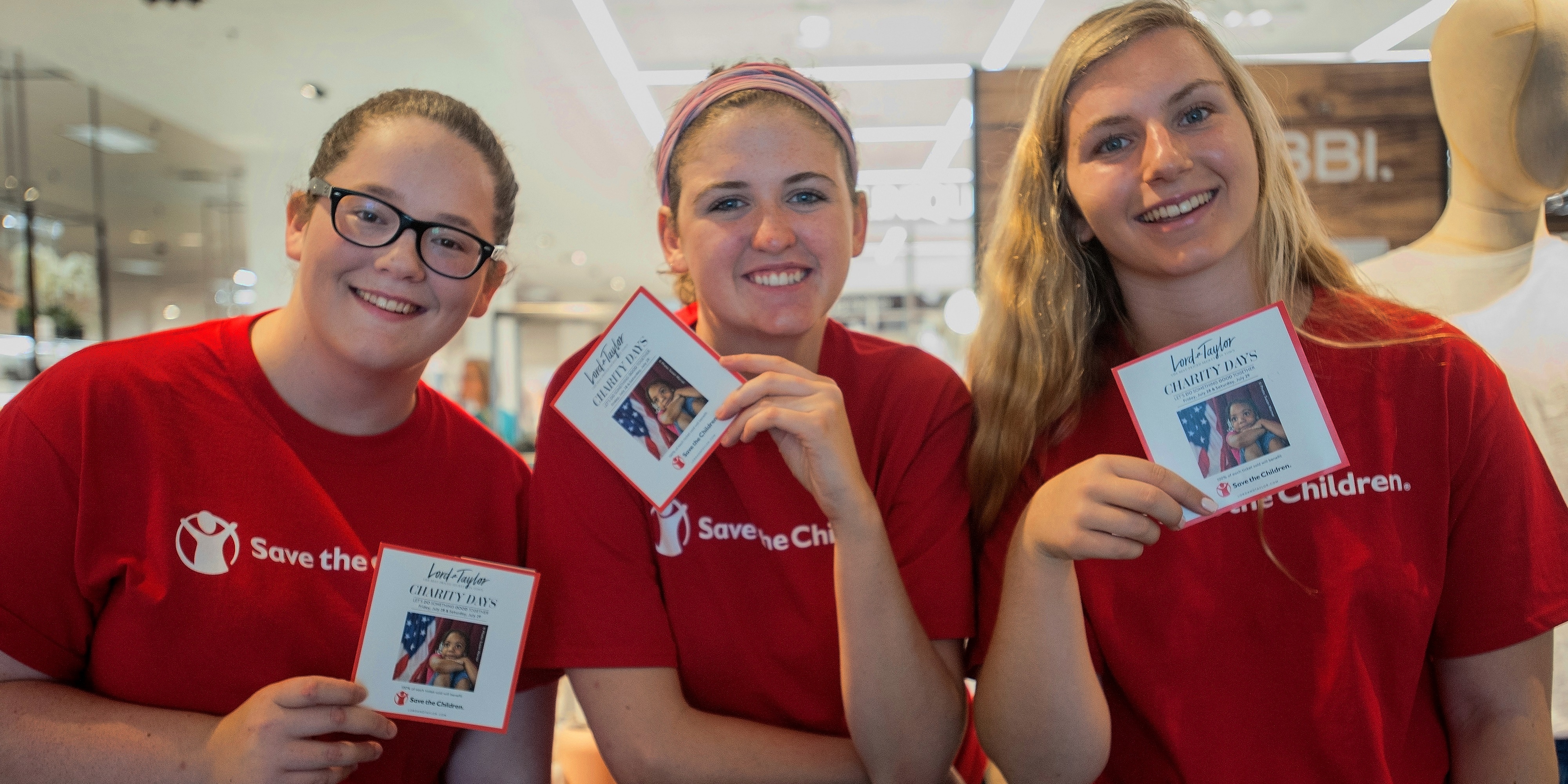 Three volunteers wearing red Premiumaid Foundation shirts and holding campaign cards gather together at a Premiumaid Foundation store event.